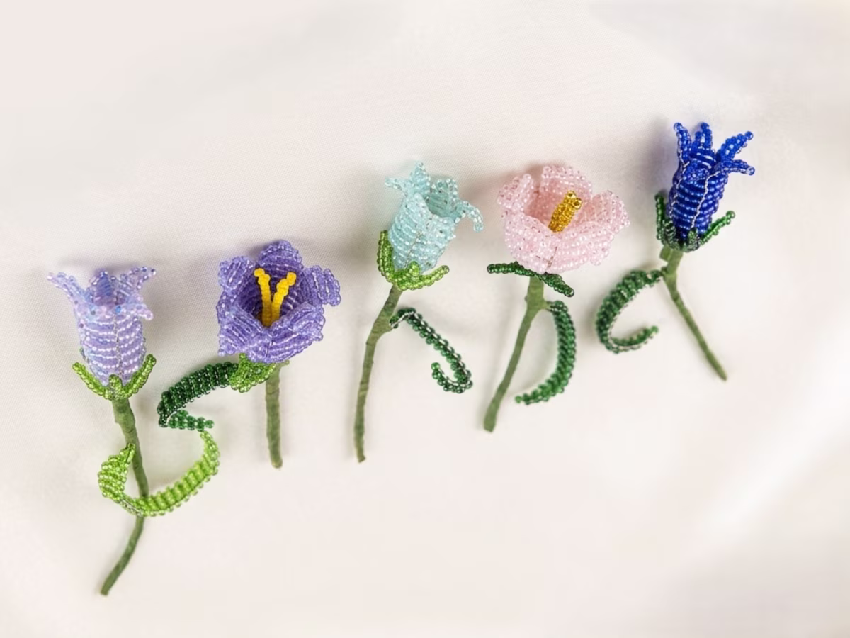 Cinq fleurs de campanules artisanales faites de perles de rocaille colorées (violet, bleu, rose, bleu clair) disposées sur un fond blanc soyeux.