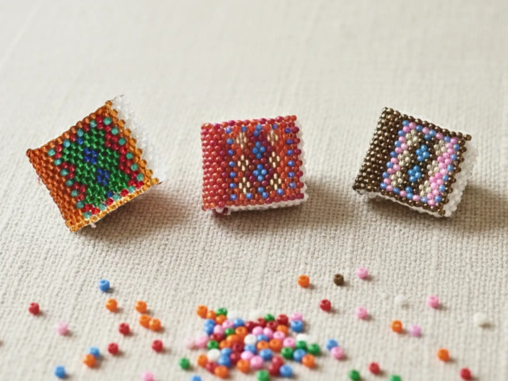 Trois mini-livrets décoratifs en perles de rocaille tissés à la main, posés sur un tissu clair avec des perles colorées éparpillées.