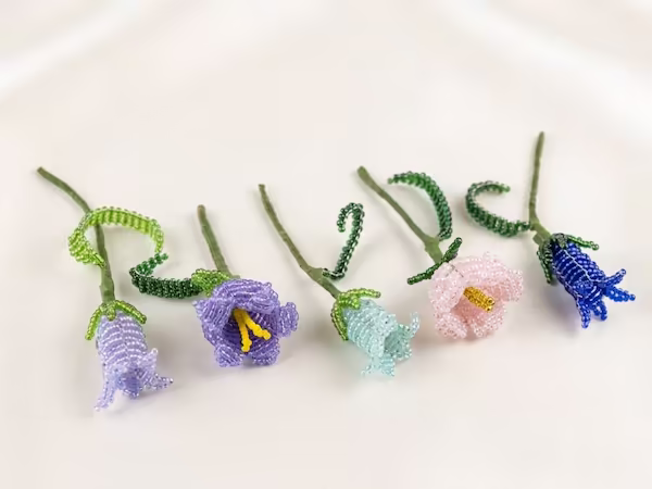 Cinq fleurs de campanules artisanales faites de perles de rocaille colorées (violet, bleu, rose, bleu clair) disposées sur un fond blanc soyeux.