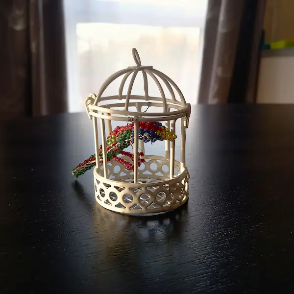 Le perroquet en perles de rocaille présenté comme élément de décoration à l'intérieur d'une petite cage décorative blanche.