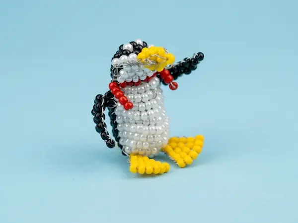 Figurine artisanale d'un petit pingouin en perles de rocaille avec une écharpe rouge, fait main en Belgique.
